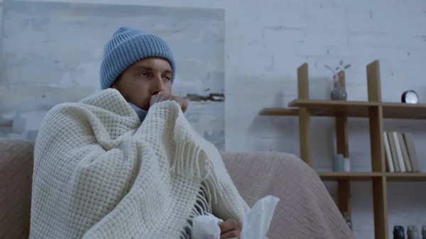 Ill man coughing while sitting on sofa in warm blanket and hat — Foto stock