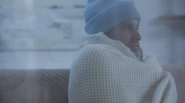 Hombre enfermo en sombrero caliente y manta sentado en el sofá en casa - foto de stock