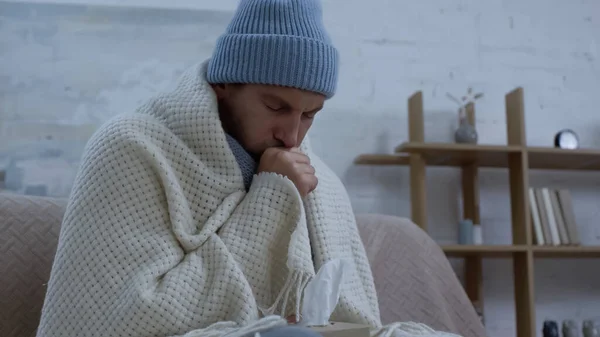 Ill man in warm hat and blanket coughing on couch at home — Photo de stock