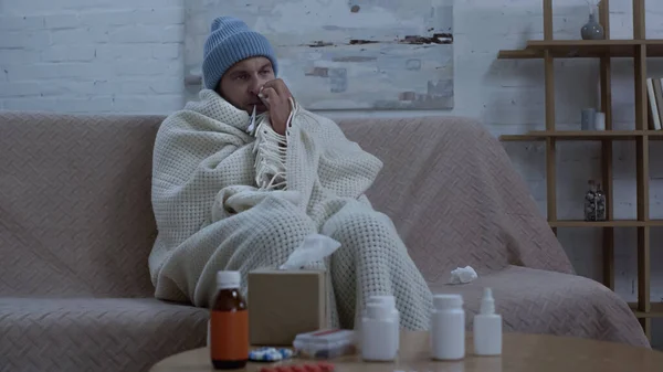 Diseased man wiping nose with paper napkin while sitting on couch in warm hat and blanket near table with medication — стоковое фото