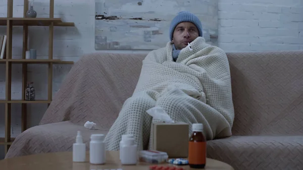 Ill man measuring temperature while sitting on couch in warm beanie and blanket near table with medication — стоковое фото
