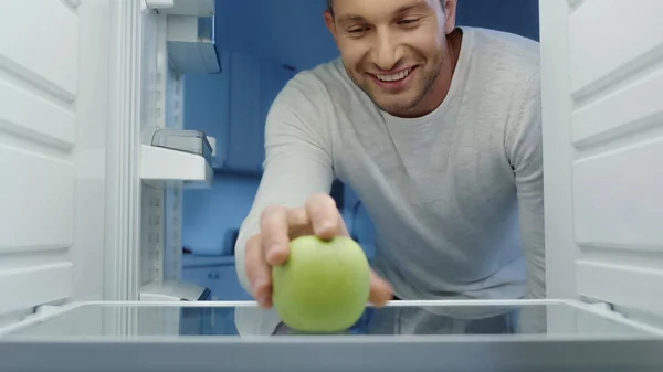 Smiling man taking green apple from fridge in kitchen — Stock Photo