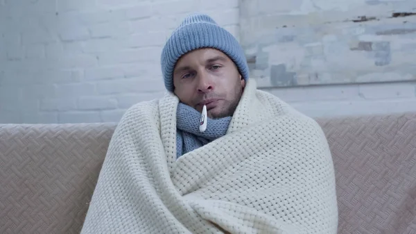 Diseased man measuring temperature while sitting on couch in warm beanie and blanket — стоковое фото