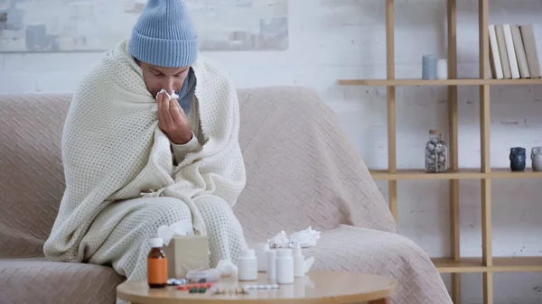 Ill man wiping nose with paper napkin while sitting in warm blanket and hat near medicaments on table — Fotografia de Stock