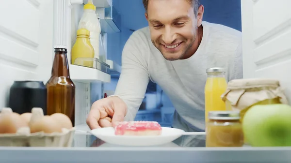 Homme heureux prenant délicieux beignet du réfrigérateur près des boissons, oeufs, bouteille et bocaux avec de la nourriture — Photo de stock