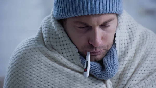 Ill man in warm blanket and beanie measuring temperature with thermometer in mouth — Stock Photo