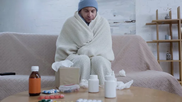 Homme malade dans un chapeau chaud et couverture assis sur le canapé près de la table avec des médicaments — Photo de stock