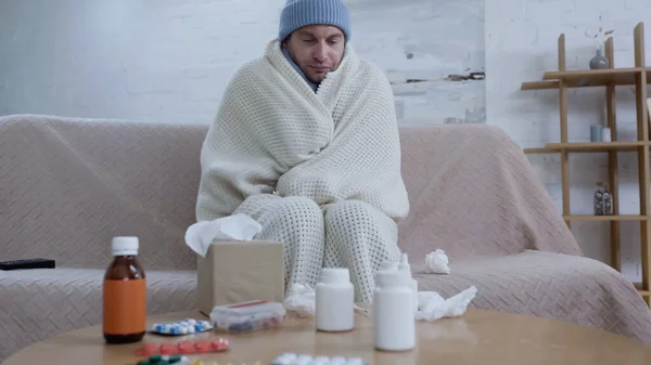 Diseased man in warm hat and blanket sitting on couch near table with medication — Fotografia de Stock