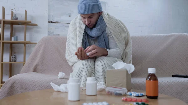 Sick man in warm blanket and hat holding pills container near table with medicaments — стоковое фото