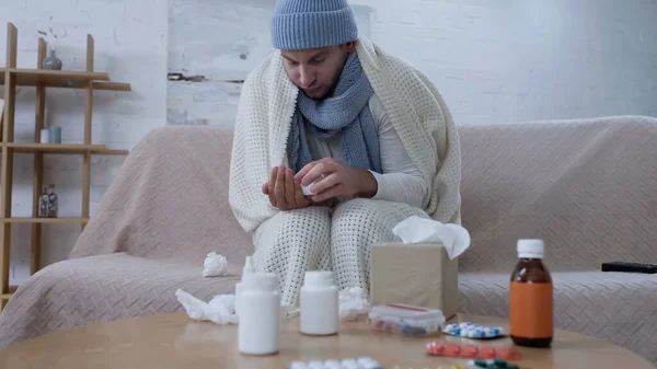 Sick man in warm blanket and hat holding pills container near table with medicaments — Foto stock