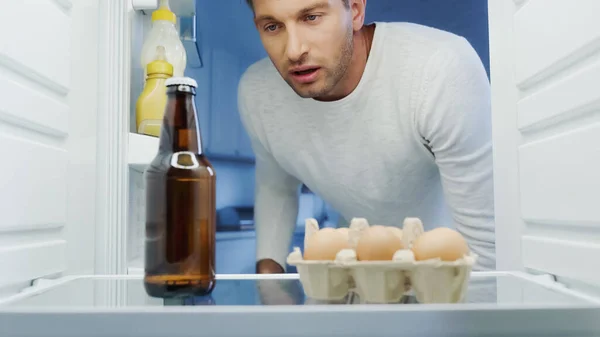Homme épuisé regardant une bouteille de bière au réfrigérateur près des œufs et des sauces — Photo de stock