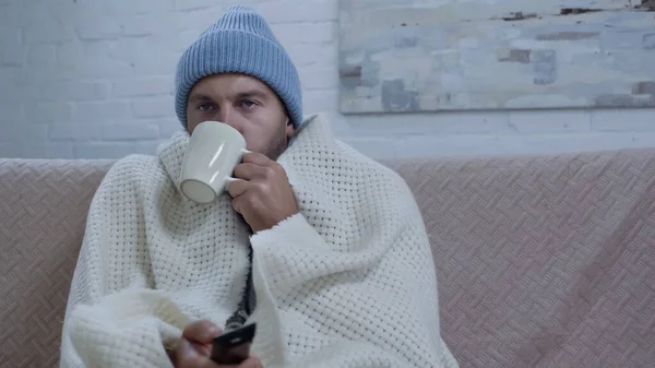 Sick man drinking warm beverage and watching tv while sitting on sofa in beanie and blanket — Stockfoto