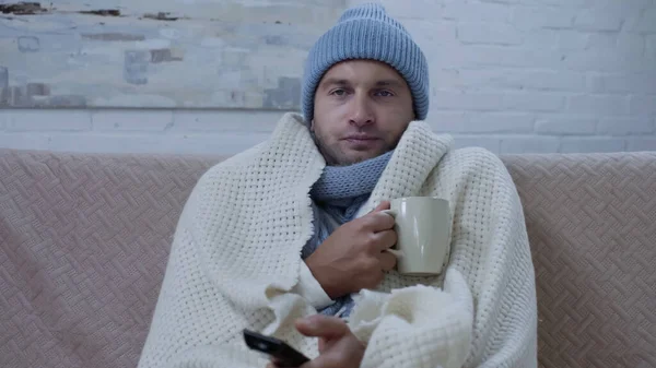 Sick man in warm blanket and hat sitting on couch with cup of tea and watching tv — Stockfoto