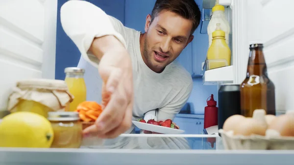 Hombre alegre tomando delicioso cupcake y fresas de la nevera con bebidas, huevos y recipientes con alimentos - foto de stock