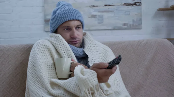 Homme malade avec tasse de thé et télécommande tv assis sur le canapé dans un chapeau chaud et couverture — Photo de stock