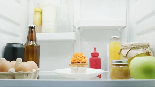 Tasty cupcake near eggs, bottle with drinks, jars with honey and fruit puree in fridge — Fotografia de Stock