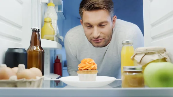 Man looking at delicious cupcake near drinks, eggs and sauces in fridge — Foto stock