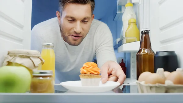 Pleased man taking plate with delicious cupcake from fridge with drinks, eggs, jars and bottles with sauces — Fotografia de Stock