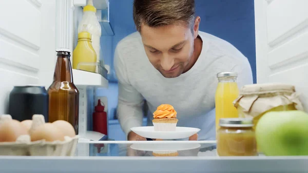 Smiling man taking cupcake from refrigerator near beverages, eggs, jars and containers with food — Stockfoto