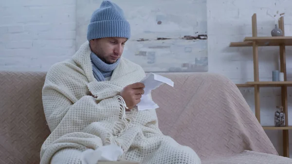 Ill man in warm beanie and blanket reading medication instruction on couch at home — стоковое фото