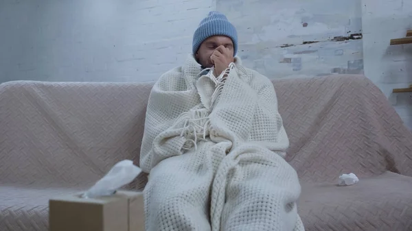 Sick man sneezing in paper napkin while sitting on sofa in warm beanie and blanket — Photo de stock