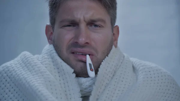 Displeased sick man with thermometer in mouth measuring temperature at home - foto de stock