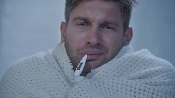 Upset diseased man in blanket looking at camera while measuring temperature with thermometer in mouth - foto de stock