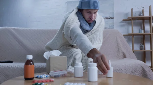 Ill man in warm blanket and hat reaching nasal spray while sitting on sofa near medicaments — Stockfoto