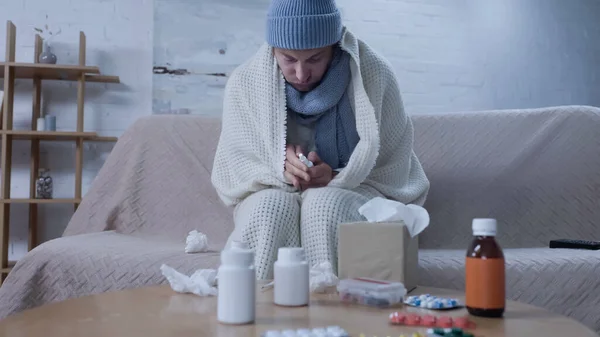 Sick man in warm hat, scarf and blanket holding nasal spray near table with medication and paper napkins — Stockfoto