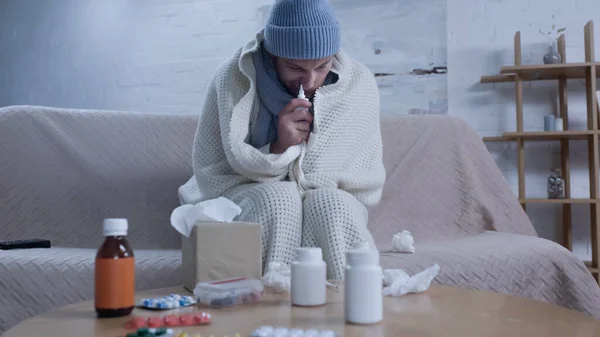 Diseased man in warm blanket and beanie using nasal spray while suffering from runny nose near table with medication — Fotografia de Stock