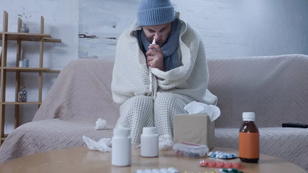 Ill man using nasal spray while sitting in warm beanie and blanket near medicaments and crumpled paper napkins — Fotografia de Stock