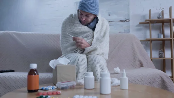 Ill man in warm hat and blanket looking at thermometer near table with medication and paper napkins — стоковое фото