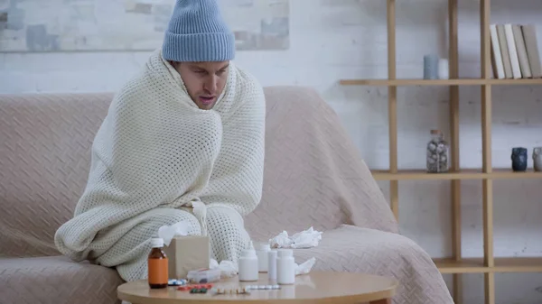 Sick man sitting on couch in warm blanket and hat near paper napkins and medicaments on table — Foto stock