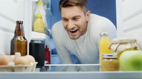 Homme excité regardant dans le réfrigérateur avec des boissons, des œufs, des pots et des bouteilles avec des sauces — Photo de stock