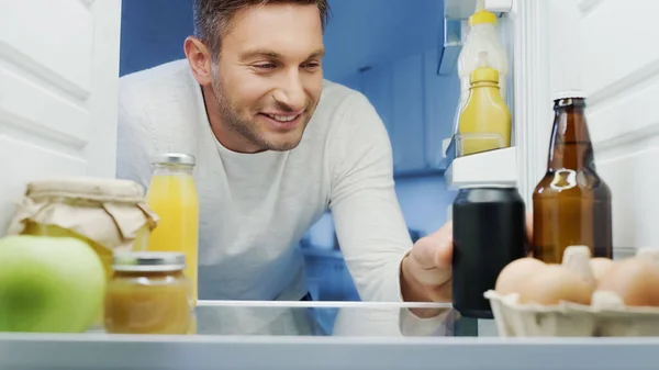 Happy man taking soda from fridge near bottle of beer, orange juice, eggs, and jars with food — Foto stock