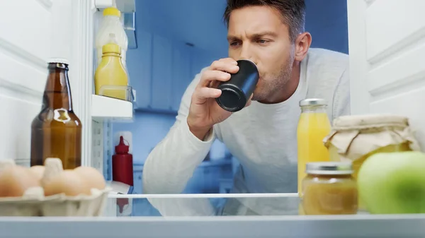 Man drinking soda from metal can near fridge with beverages, eggs and bottles with sauces — Stock Photo