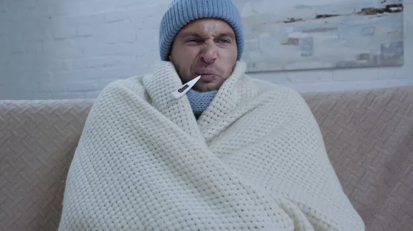 Незадоволений хворий чоловік у теплому капелюсі та ковдрі вимірювальна температура з термометром у роті — стокове фото