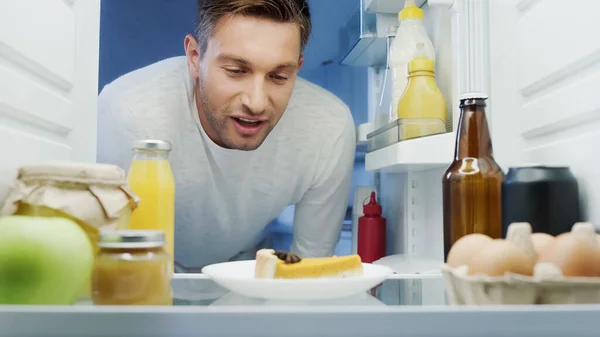 Pleased man looking at delicious cake in fridge near beverages, eggs and sauces — Photo de stock
