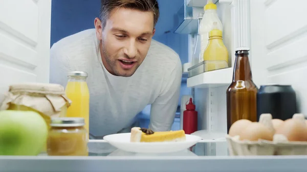 Pleased man looking at delicious cake in fridge near beverages, eggs and sauces — Stock Photo