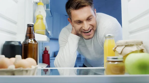 Cheerful man looking into refrigerator with fresh orange juice, beer, eggs and bottles with sauces — Fotografia de Stock