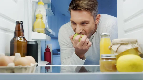 Man eating ripe apple near fridge with eggs, beverages, containers and jars with food — Stock Photo