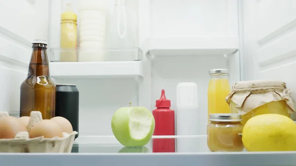 Manzana mordida, botellas con bebidas y salsas, limón, miel y puré de frutas en nevera - foto de stock