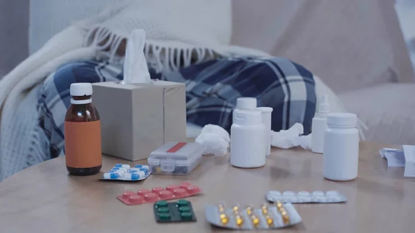 Cropped view of sick man sitting on couch under warm blanket near table with medicaments and paper napkins - foto de stock