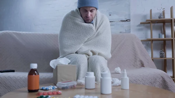 Diseased man in warm blanket and hat sitting with closed eyes near table with medication and paper napkins — Stock Photo
