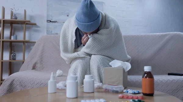 Ill man sneezing in paper napkin while sitting on couch near medicaments on table — Stock Photo