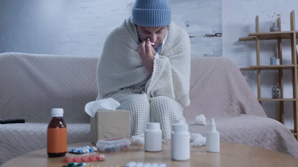 Hombre en manta caliente y gorro que sufre de secreción nasal cerca de la mesa con medicamentos - foto de stock