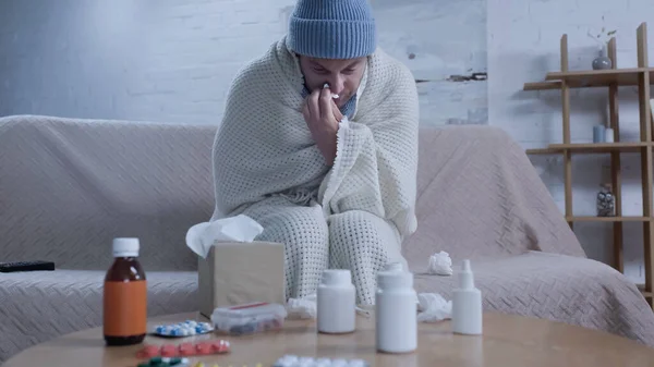 Man in warm blanket and beanie suffering from runny nose near table with medication — Stock Photo