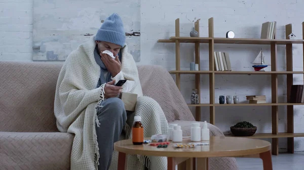 Sick man in warm beanie and hat clicking tv channels while suffering from runny nose near medication on table — Stockfoto