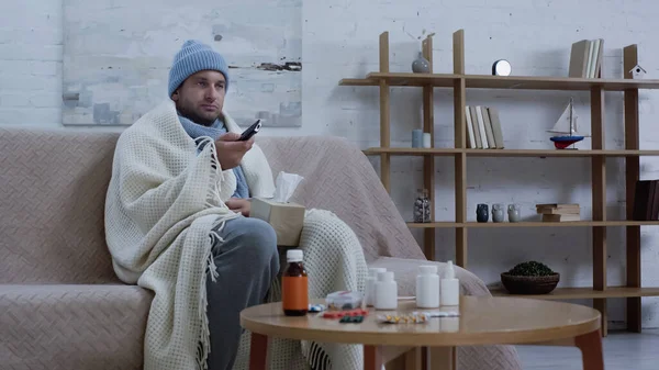 Sick man in warm beanie and blanket clicking tv channels near medicaments on table — Photo de stock