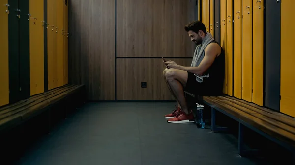 Deportista sentado en el banco y charlando mientras sostiene el teléfono inteligente en el vestuario - foto de stock